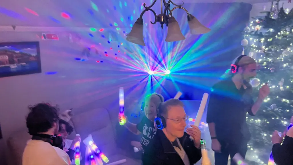 People of all ages in silent disco headphones waving glow sticks under disco lights, beside a Christmas tree.
